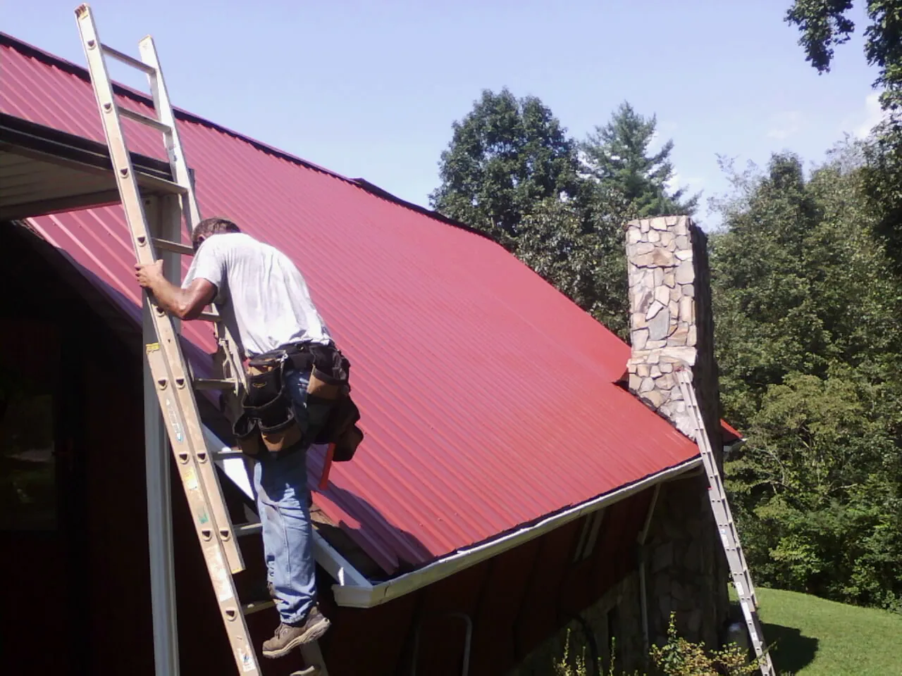 Roofer on a ladder beside a red metal roof and stone chimney — Soffit & Fascia Repair by Roofing Brothers in Islandia